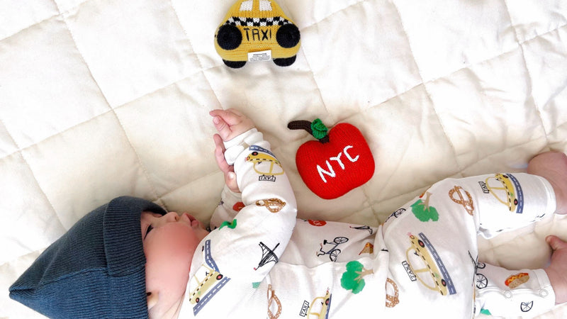 Baby wearing NYC print romper lying with taxi & apple rattles.