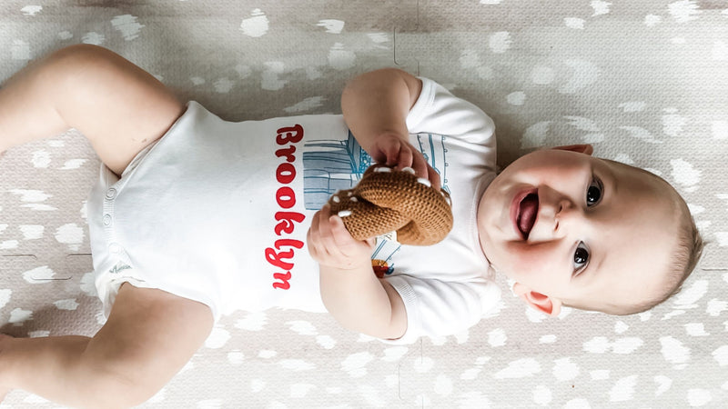 Baby in 'Brooklyn' onesie playing with pretzel rattle toy.