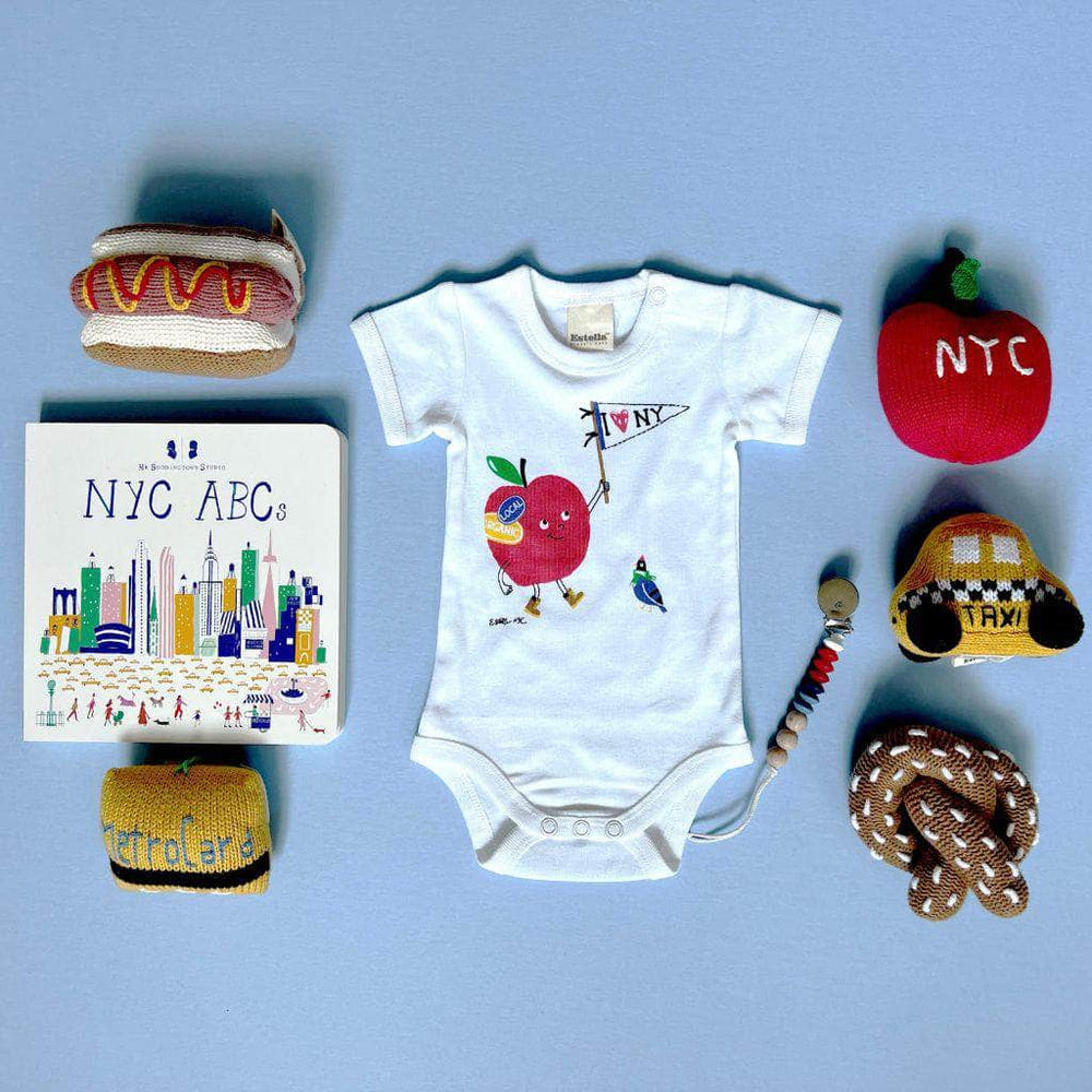 "I Love NY" baby gift set which includes cream and yellow striped organic onesie with red apple graphic, brown pretzel rattle, yellow taxi rattle, red apple rattle with "NYC" embroidered in white, hot dog rattle, yellow metrocard rattle, NYC ABCs baby board book and beaded infant pacifier clip in red, blue, white and natural wood.