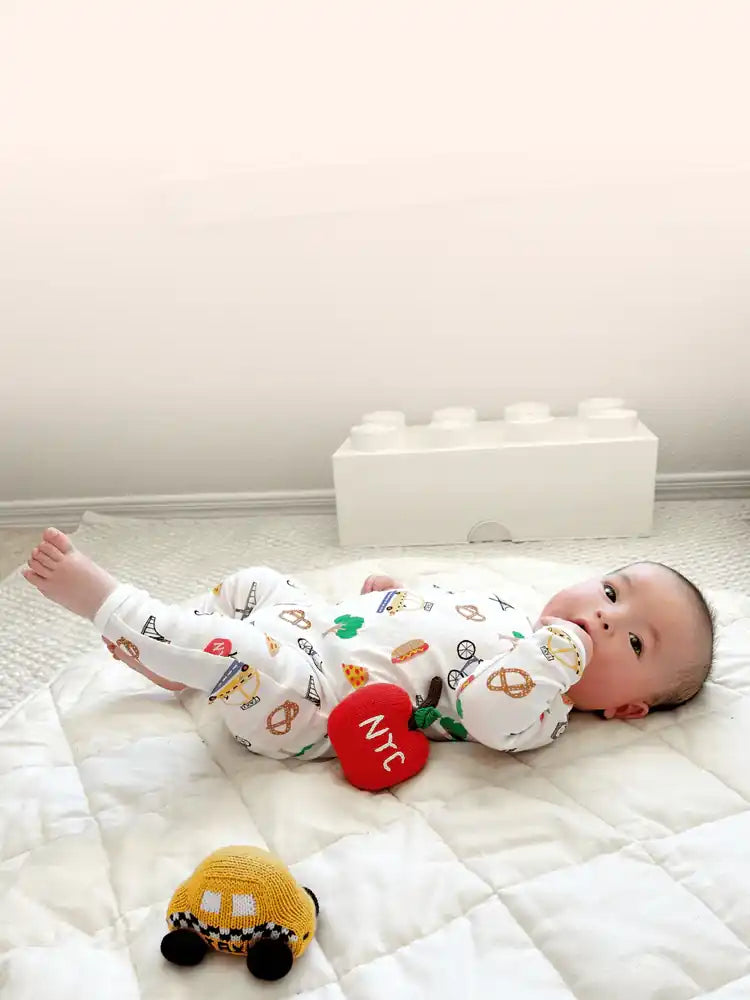 Baby lying on a white mattress with toys, wearing a patterned onesie.