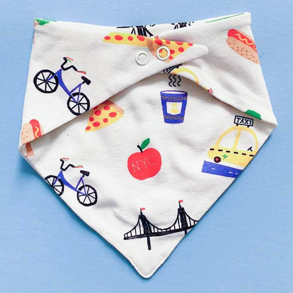 Back of the Triangular baby bib that snaps in the back. Features a white background with small images of pretzels, apples, hotdogs, pizza, trees, coffee, taxis, bikes and bridges. 