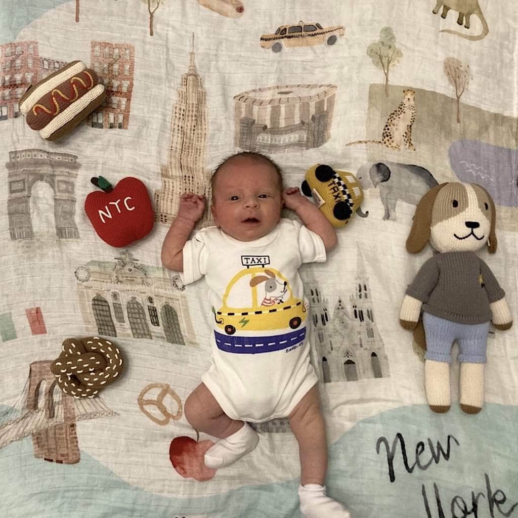 Newborn baby wearing the white taxi onesie with yellow taxi, surrounded by Estella NYC rattles and toys.