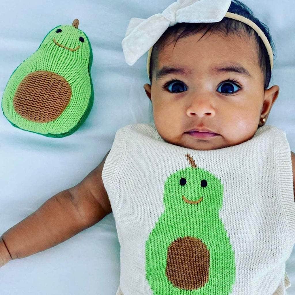 Baby girl wearing the cream-colored knitted Avocado sunsuit with bright green, smiling avocado intarsia in the center.