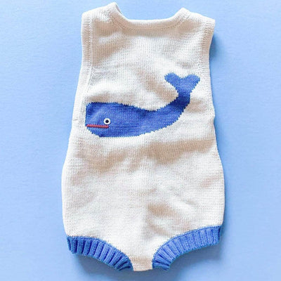 Cream-colored whale baby sunsuit with graphic, happy whale intarsia in the center. Leg holes have blue ribbed binding to match whale. Photographed on a vibrant blue background.