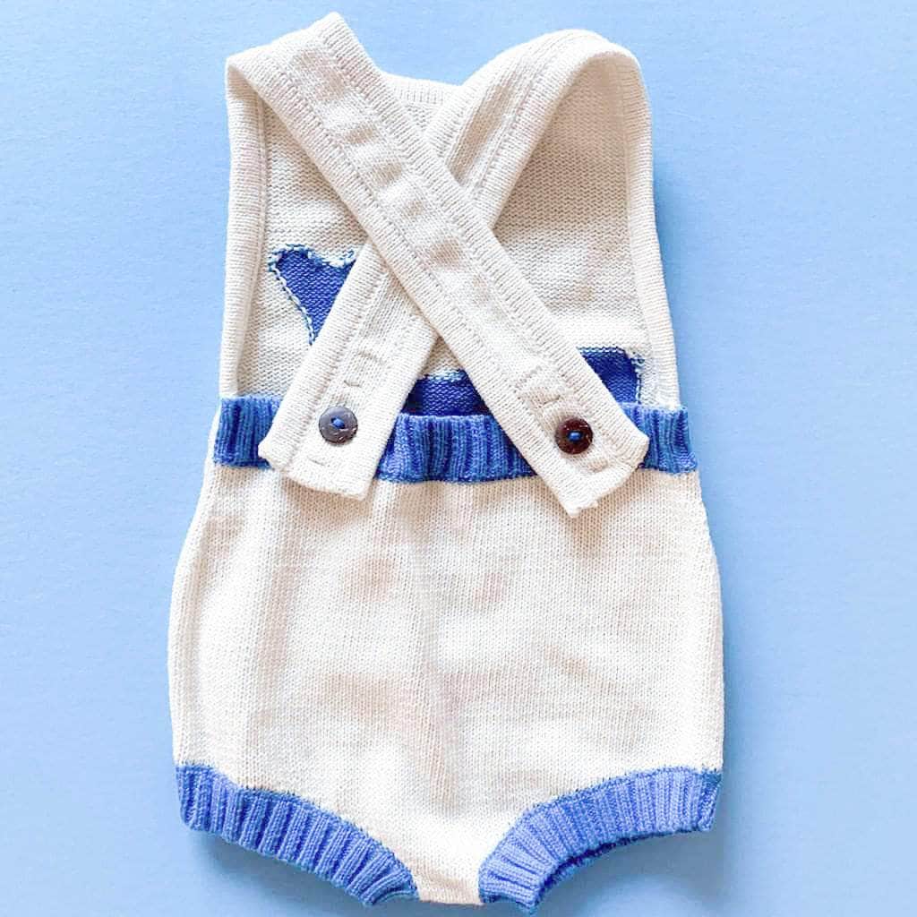 Back of baby whale sunsuit shows cris-crossed straps with dark brown wood buttons and three button holes on each strap so straps can be made longer or shorter to fit baby as he/she grows. Back waist band is ribbed and blue . Photographed on a vivid blue background.