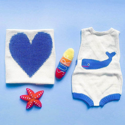 Surfside baby gift set features cream whale knitted summer romper with blue whale, ribbed cream heart lovey with heart, red starfish rattle and multi-colored surfboard rattle. Photographed on a bright blue background.