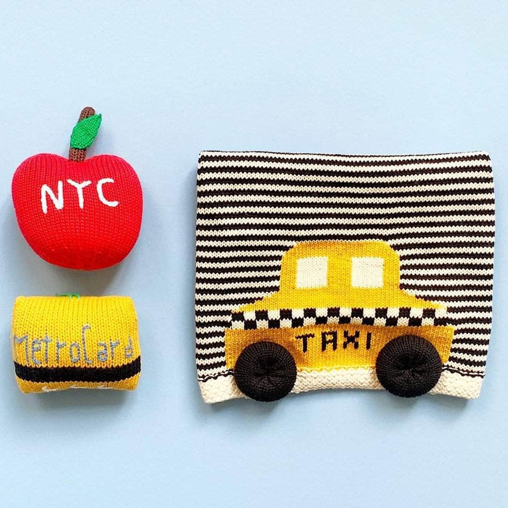 Image of a red apple baby rattle, a yellow metrocard baby rattle and a black and white striped baby blanket with a yellow taxi. All photographed on a soft blue background.