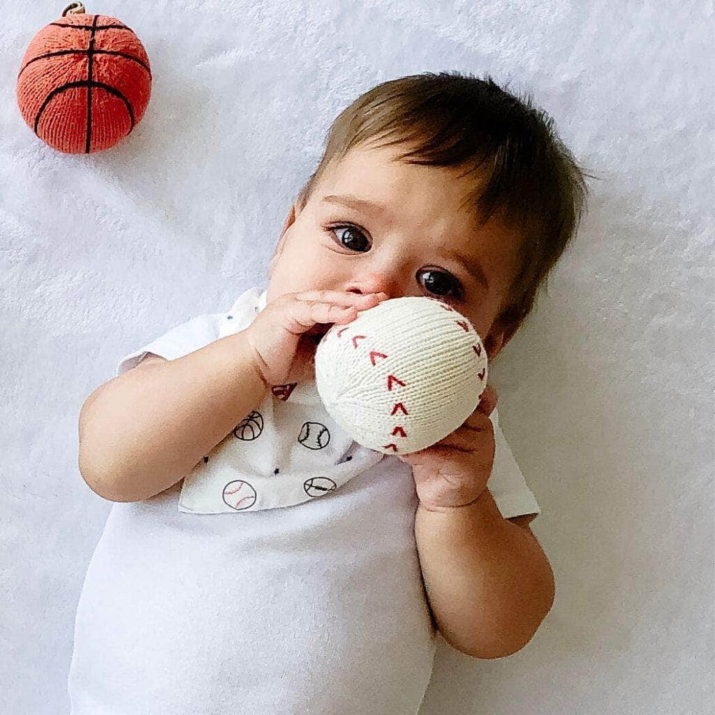 baby hold a baseball rattle and basketball rattle next to him.