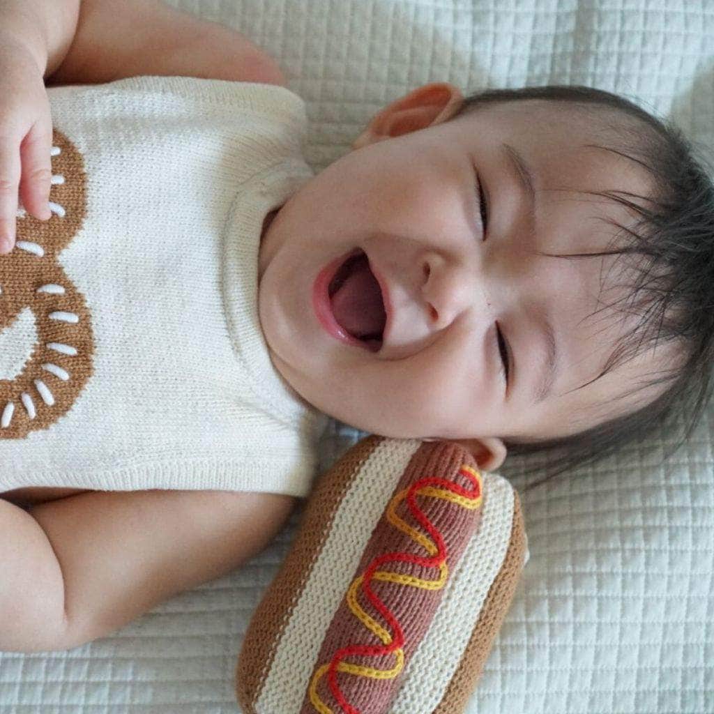 organic baby rattle toy hot dog next to a baby wearing the sleeveless pretzel romper. 