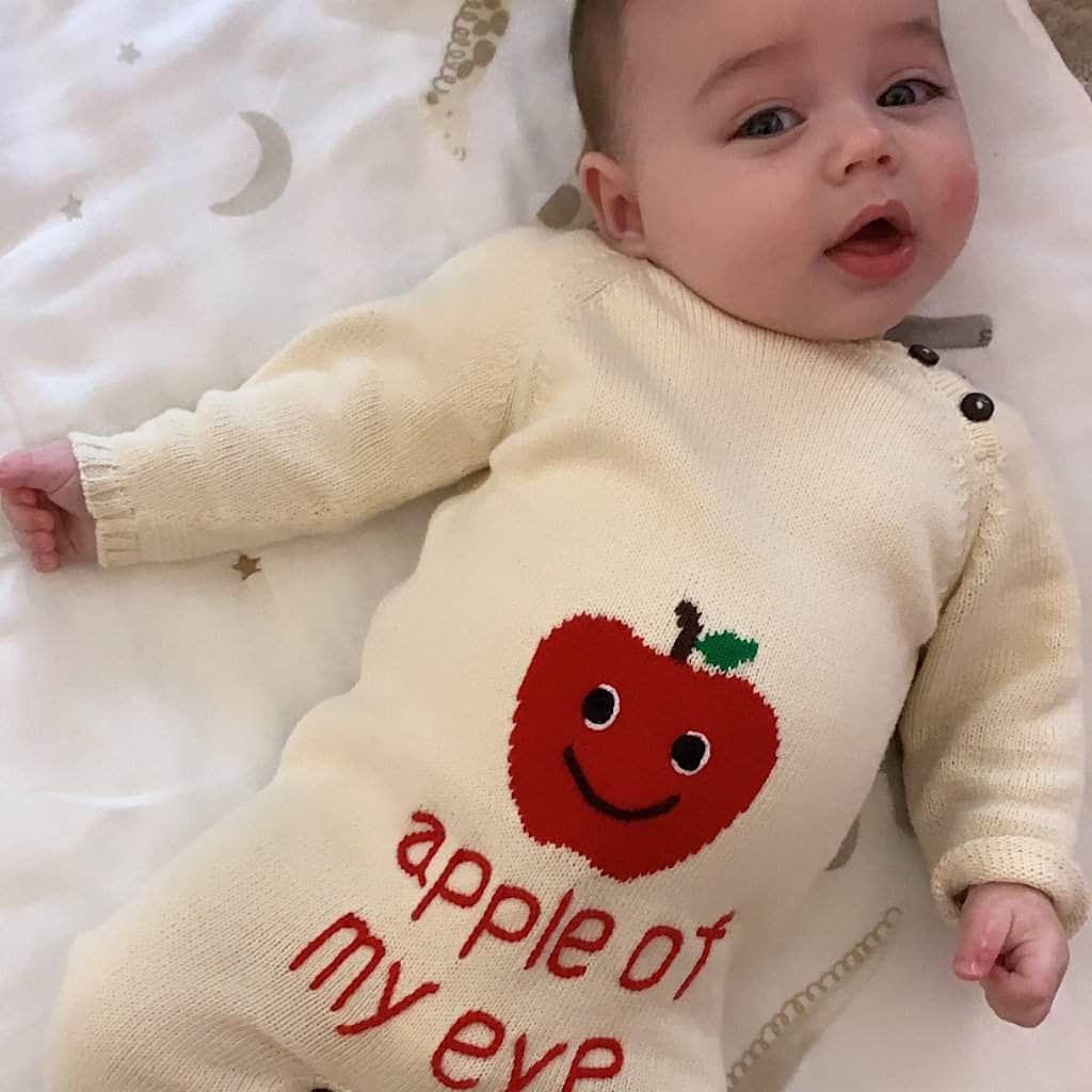 Sweet baby, lying on a quilt, wearing the cream romper  with red apple intarsia and red "apple of my eye" embroidery.