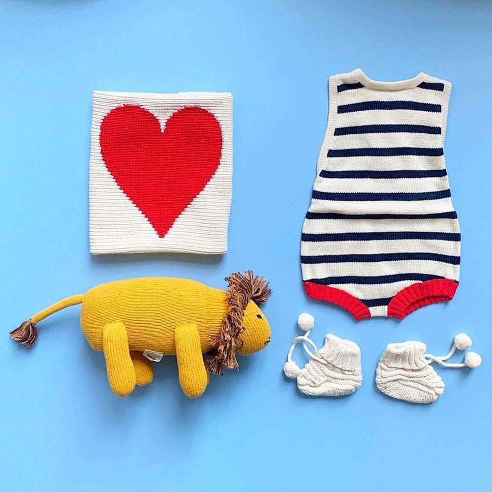 Photo of navy blue and cream striped summer onesie, yellow stuffed lion toy, cream booties and cream baby lovey with red heart. Photographed on a blue background.