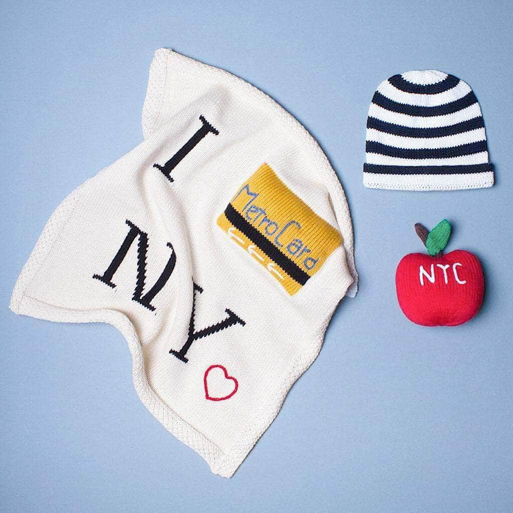 Photograph of cream "I Metro NY" lovey blanket with small red heart embroidered on the bottom right corner of the lovey, red apple rattle with the letters "NYC" embroidered on top in white, and a black and cream striped baby hat. Photographed on a soft gray background.