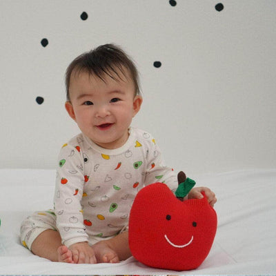 Baby, sitting on a bed and smiling, grasping the stuffed organic apple toy. The apple is red with a black stem, green leaf and large, happy face.