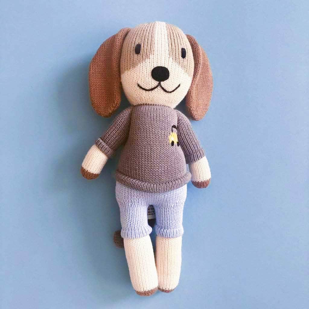 Cream and brown knitted dog doll, Frank. He has brown spots around his eyes and floppy brown ears, and smiles broadly. He wears a gray sweater with an embroidered taxi at his breast. He wears blue knitted shorts and has cream arms, face and legs. Shown on a blue background. 