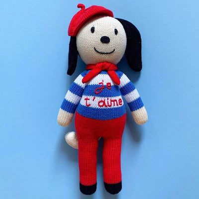 Picture of cream-colored knit dog doll with striped blue sweater, red pants, red beret and red scarf.  The sweater is hand-embroidered with the words "je t'aime." Photographed on a vivd blue background.