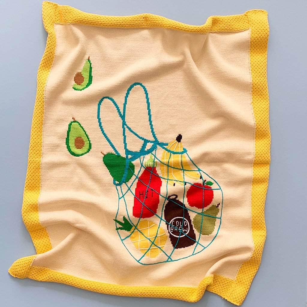 Hand-knitted baby blanket in cream with bright yellow border. The blanket features intarsia images of pineapples, bananas, avocados, hot sauce, apple, pear and a cold brew coffee wrapped up in a top-stitched string grocery bag.  The blanket is bright and colorful and photographed on a blue background.