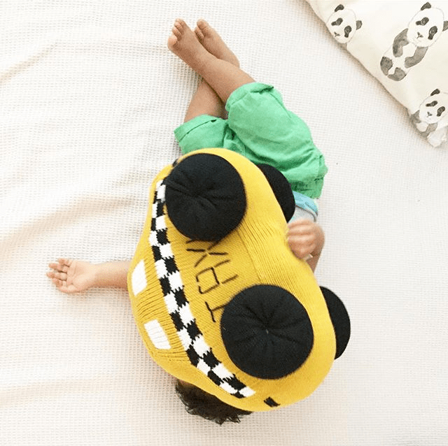 Toddler, lying on a bed, hiding behind the large taxi stuffed toy. All that shows behind the yellow taxi are the boy's legs, feet and hands.