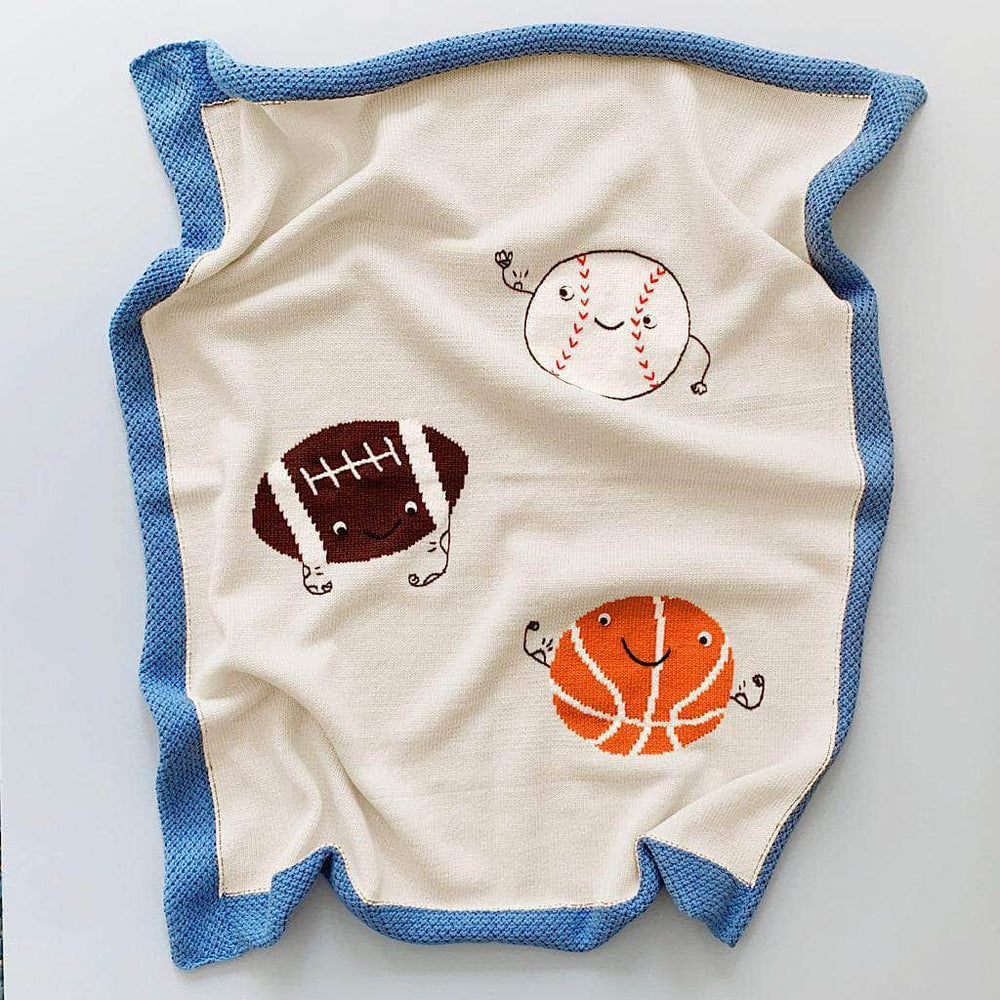 Photo of a cream knitted blanket with images of an orange basketball, a white baseball, and a brown football. All sports balls have smiling faces and flexing their muscles. Blanket has a blue border and is photographed on a blue background.
