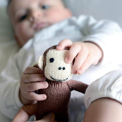 Animal baby toy monkey. This rattle is organic, soft & perfect for little hands. Brown with a white smiling face, it is shown held by a baby.