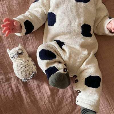 Baby wearing a cream and navy knitted  Estella romper, lying on a bed next to the cream and gray Estella owl  rattle